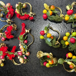 May include: Ten gold-toned curtain tiebacks with red, green, and yellow berry-like decorations. The tiebacks are decorated with faux greenery and berries.