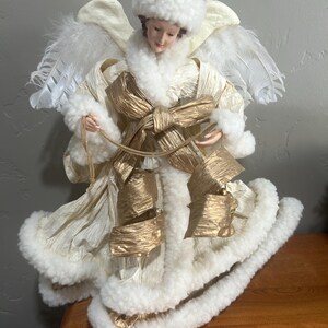 May include: A decorative angel figurine with a cream-colored dress, white feathered wings, and a matching hat. The angel has a gold bow and trim, and is designed to be a tree topper.