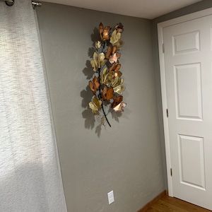 May include: A large metal wall sculpture featuring a stylized branch with leaves in shades of gold, copper, and brown. The sculpture is mounted on a light gray wall.