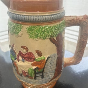 May include: A ceramic pitcher with a brown rim and handle, featuring a detailed scene of people gathered under trees. The pitcher has a textured design around the middle and a spout for pouring.