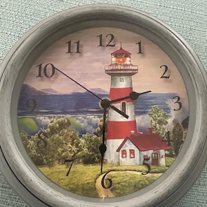 May include: Round wall clock with a lighthouse scene. The clock face features a red and white striped lighthouse, a small house, and a coastal landscape. The clock has a gray frame and black hands.