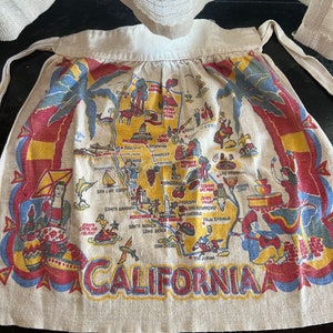 May include: A beige linen kitchen apron with a map of California printed on it. The map features various cities and landmarks, including Los Angeles, San Francisco, San Diego, and Palm Springs. The map is surrounded by a red, blue, and yellow border.