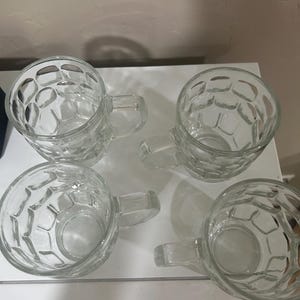 May include: Four clear glass beer mugs with a textured, honeycomb-like pattern. Each mug has a handle and a rounded base. The mugs are arranged on a white surface, perfect for serving beer or other beverages.