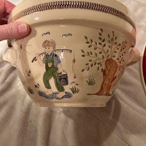 May include: A beige ceramic bowl with a decorative rim and handles. The bowl features a painted scene of a child fishing, a tree, and birds. The child is wearing green overalls and carrying a bucket. The bowl has a rustic, vintage aesthetic.
