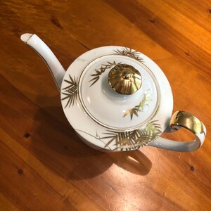 May include: White teapot with a gold lid and gold leaf design. The teapot has a spout and a handle.