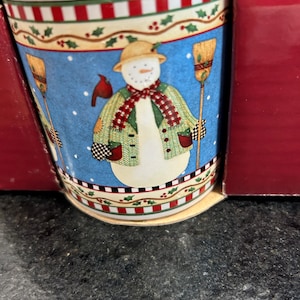 May include: A white ceramic mug with a snowman design. The snowman is wearing a hat, scarf, and gloves. The mug has a blue background with red and white stripes around the top.