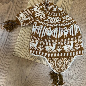 May include: A brown and white knitted hat with a llama pattern and the word "Peru" repeated on the brim. The hat has a long tassel on the top and a shorter tassel on the bottom.