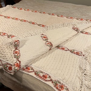 May include: A crocheted throw blanket with a white background and a floral pattern in shades of orange and green. The blanket has a fringe border.