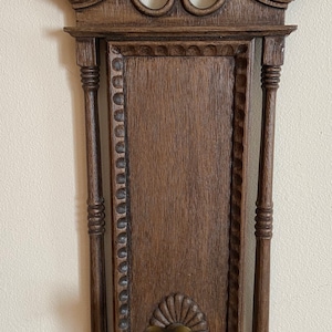 May include: A dark brown wooden wall sconce with ornate carvings and a brass candle holder. The sconce has a rectangular frame with a decorative border and a scalloped design at the bottom.
