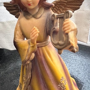May include: A figurine of an angel with brown hair and a yellow dress holding a lyre. The angel has wings and is wearing a gold belt.