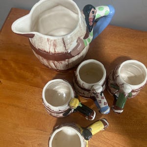May include: A vintage ceramic pitcher and matching mugs set. The pitcher has a barrel design with a handle featuring a Native American figure. The mugs also have a barrel design with handles featuring Native American figures. The set is painted in shades of brown, white, blue, green, and yellow.