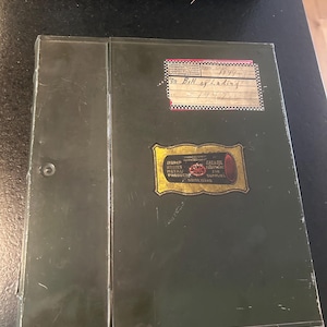 May include: An olive green metal document box with a hinged lid. A rectangular label at the top reads "Bill of Lading 1947". A decorative gold-bordered label in the center features the text "DUMP BODIES METAL PRODUCTS LOMC GAS & OIL EQUIPMENT AND SUPPLIES BOISE IDAHO".