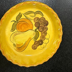May include: Yellow ceramic pie plate with a hand-painted design of a pear, peach, and grapes. The plate has a scalloped edge.