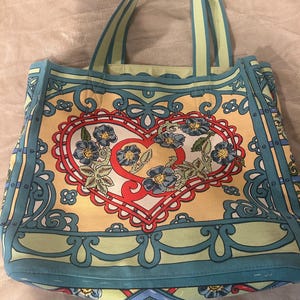 May include: A colorful tote bag with a heart-shaped floral design. The bag features a yellow background with a red heart, blue flowers, and green leaves. The handles and trim are light blue and green.