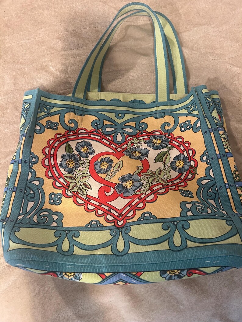 May include: A colorful tote bag with a heart-shaped floral design. The bag features a yellow background with a red heart, blue flowers, and green leaves. The handles and trim are light blue and green.