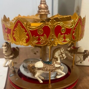 May include: A rotating carousel candle holder featuring white horses with gold saddles and bridles. The structure has a red and gold ornate canopy with a central spire and a circular base designed to hold a small candle in the center.