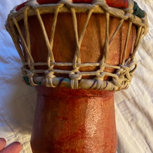 May include: A small, red wooden drum with a white rope lacing pattern. The drum has a red and white striped rim and a blue and white striped base.