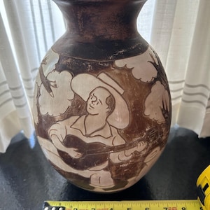May include: A brown ceramic vase with a detailed relief carving of a musician playing a guitar. The vase is approximately 8 inches tall and features a dark brown rim and base, with lighter brown and white accents.