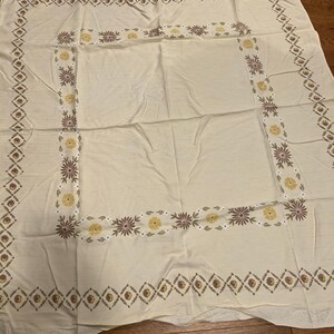 May include: Cream-colored tablecloth with a floral and geometric border design. The border features alternating diamond and floral patterns in shades of brown, yellow, and white. The tablecloth is rectangular and appears to be made of a soft fabric.