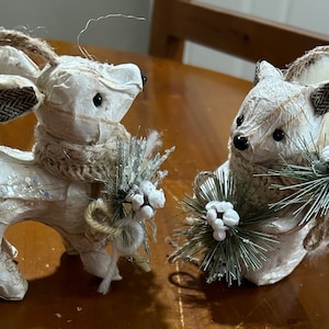 May include: Two white, paper mache Christmas ornaments, a deer and a squirrel, both wearing a brown scarf and holding a small pine sprig with white berries.