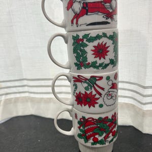 May include: A stack of four white ceramic Christmas mugs. Each mug features a different festive design: reindeer, holly and poinsettias, Santa Claus, and Christmas bells. The mugs have curved handles and are stacked on top of each other.