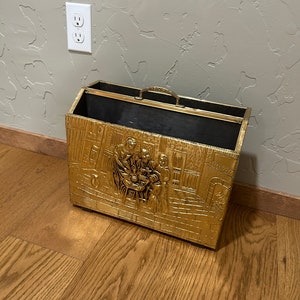 May include: A gold-colored metal log holder with an embossed design of people and a fireplace. The holder has a black interior and a handle on the top.