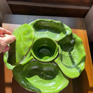 May include: A green ceramic serving dish with a central bowl and four surrounding leaf-shaped sections. The dish is a vibrant green color and has a glossy finish. The design is reminiscent of a lettuce leaf. The dish is on a wooden surface.