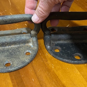 May include: Two vintage metal handles with a curved design. One is a weathered gray, the other is black. Each has a rectangular base with rounded edges and several mounting holes, suggesting they are for a trunk or chest.