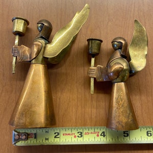 May include: Two vintage brass angel candle holders. The angels have copper-colored bodies, brass wings, and hold candle cups. The larger angel is approximately 5 inches tall, and the smaller one is about 4 inches tall. The angels are on a wooden surface.