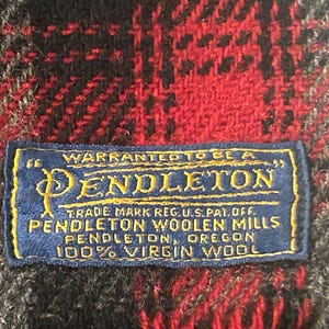 May include: A red, black, and grey plaid textile featuring a rectangular blue label with yellow text that reads: WARRANTED TO BE A PENDLETON, TRADE MARK REG. U.S. PAT. OFF., PENDLETON WOOLEN MILLS, PENDLETON, OREGON, 100% VIRGIN WOOL.