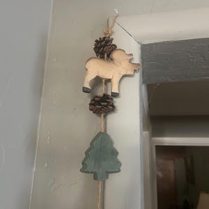 May include: A rustic wooden wall hanging featuring a carved moose, a green tree, and pine cones. The moose is light brown with black feet. The tree is painted green. The pine cones are brown and attached with twine.