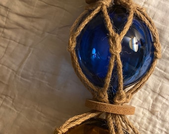 May include: Three glass fishing floats, each wrapped in natural twine, are strung together. The top float is green, the middle float is blue, and the bottom float is amber.