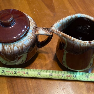 May include: A brown and white glazed ceramic creamer and sugar bowl set. The creamer has a handle and a spout. The sugar bowl has a lid. The set is on a wooden surface.