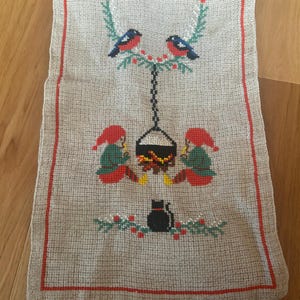 May include: A beige cross-stitch wall hanging with a red border. The design features two birds perched on a branch, two figures in red hats, a black cat, and a hanging pot over a fire. The overall theme is a folk art style.