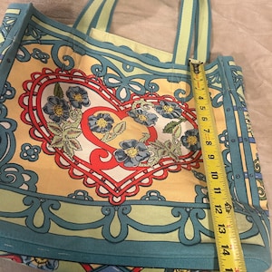 May include: A colorful tote bag with a floral heart design. The bag is approximately 15 inches tall. The bag features a yellow background with blue and red accents. The handles are light green with a blue stripe. The bag is made of a sturdy material.