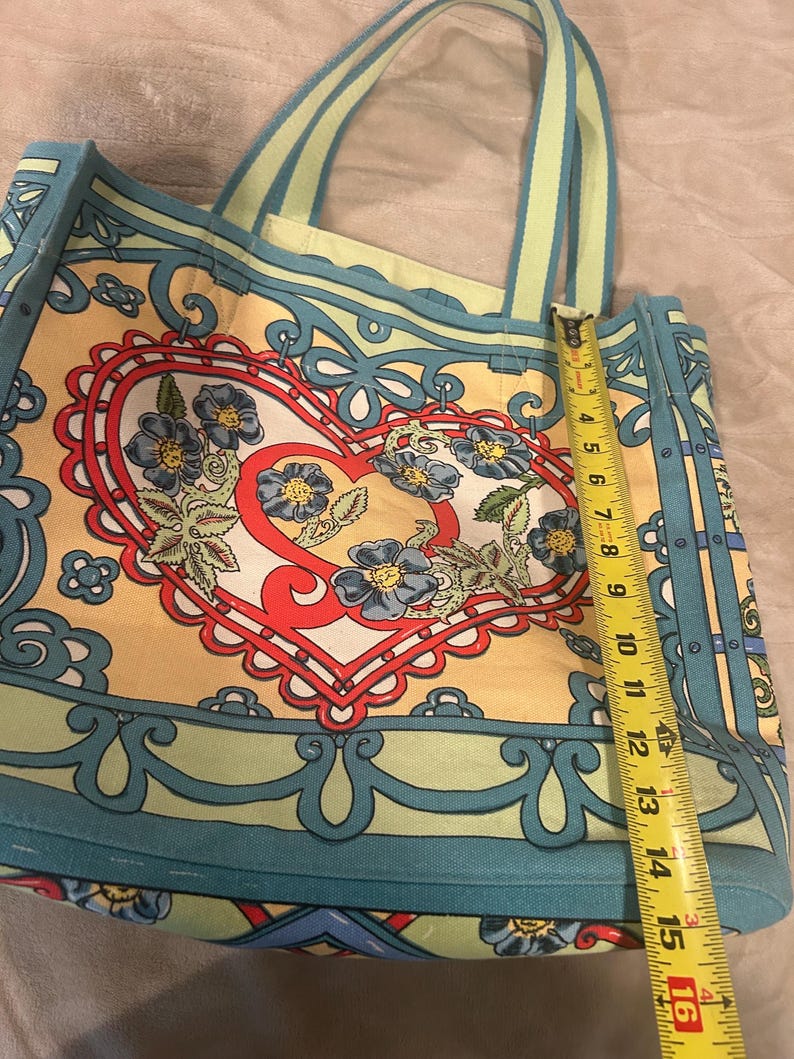 May include: A colorful tote bag with a floral heart design. The bag is approximately 15 inches tall. The bag features a yellow background with blue and red accents. The handles are light green with a blue stripe. The bag is made of a sturdy material.