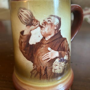 May include: Vintage ceramic mug with a brown handle and base. The mug features a detailed illustration of a monk drinking from a gourd, with a basket of grapes. The mug has a cream-colored background with a brown rim.