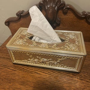 May include: A gold and brown tissue box with ornate floral designs. The box is made of plastic and has a clear plastic window on the top.