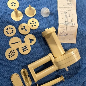 May include: A vintage cookie press machine with a beige plastic body and a crank handle. The machine is disassembled and laid out on a blue surface. The set includes a variety of cookie cutters, a nozzle, and instructions. The instructions are titled "The Cookie Machine".