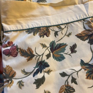May include: White cotton fabric with a floral pattern of green, brown, and pink leaves and flowers. The fabric has a cream-colored border with a thin blue trim.
