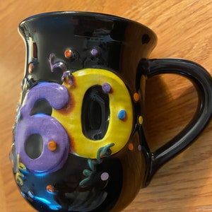 May include: Black ceramic mug with a raised 3D design of the number 60. The number is yellow with purple accents and surrounded by colorful confetti.