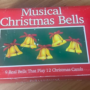 May include: A red box with the words "Musical Christmas Bells" in white. The box features an image of five yellow bells with red bows on a green background. Text on the box reads "9 Real Bells That Play 12 Christmas Carols."