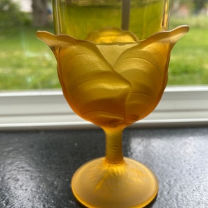 May include: A yellow glass wine goblet with a rose design. The goblet has a wide bowl and a tall stem.