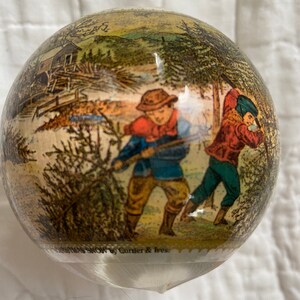 May include: A vintage glass Christmas ornament with a painted scene of two people in a snowy forest. The ornament is clear and has a white, feathery texture. The text "Christmas Snow by Currier & Ives" is printed on the bottom of the ornament.
