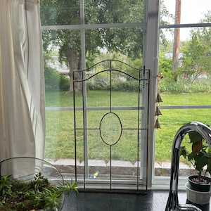 May include: A metal window grate with a decorative floral design in the center. The grate is in front of a window with a view of a green lawn and trees.