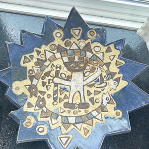 May include: A ceramic star-shaped plate with a blue glaze and a central design. The design features a figure with geometric patterns in tan, brown, and white. The plate has a textured surface and is likely handmade.
