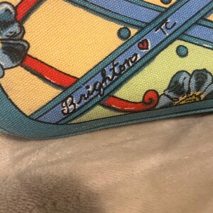 May include: Close-up of a patterned fabric with a yellow background, blue ribbons, and red accents. The word "Brighton" is written in cursive on a blue ribbon, with a heart symbol. The fabric has a teal border.