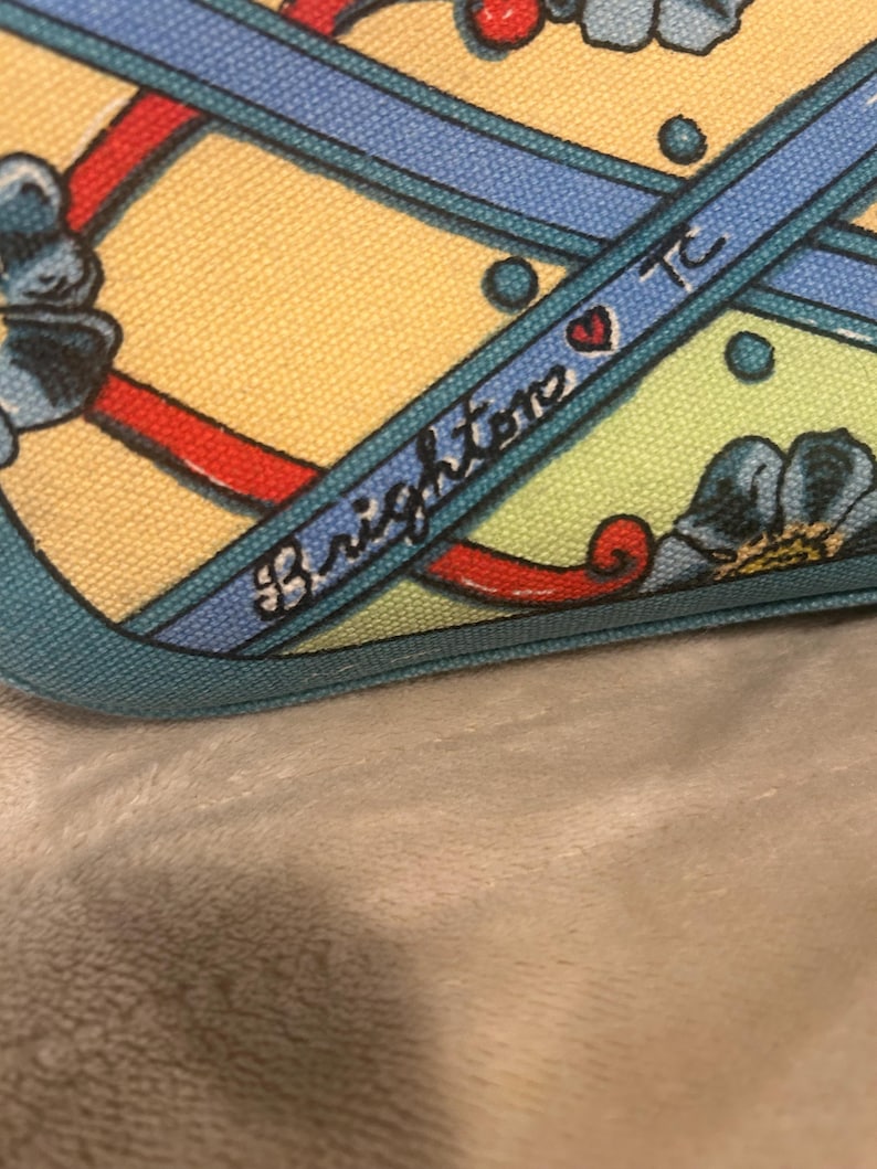 May include: Close-up of a patterned fabric with a yellow background, blue ribbons, and red accents. The word "Brighton" is written in cursive on a blue ribbon, with a heart symbol. The fabric has a teal border.