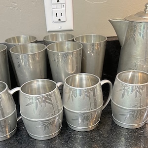 May include: A set of silver-colored mugs and a pitcher with a bamboo design. The mugs have handles and the pitcher has a spout and a lid.