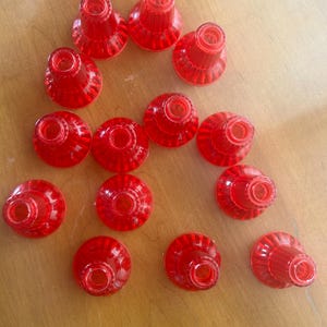 May include: A collection of thirteen translucent red plastic objects, possibly bottle stoppers or decorative elements. Each piece has a conical shape with a ridged exterior and a circular opening at the top. They are arranged on a light brown wooden surface.